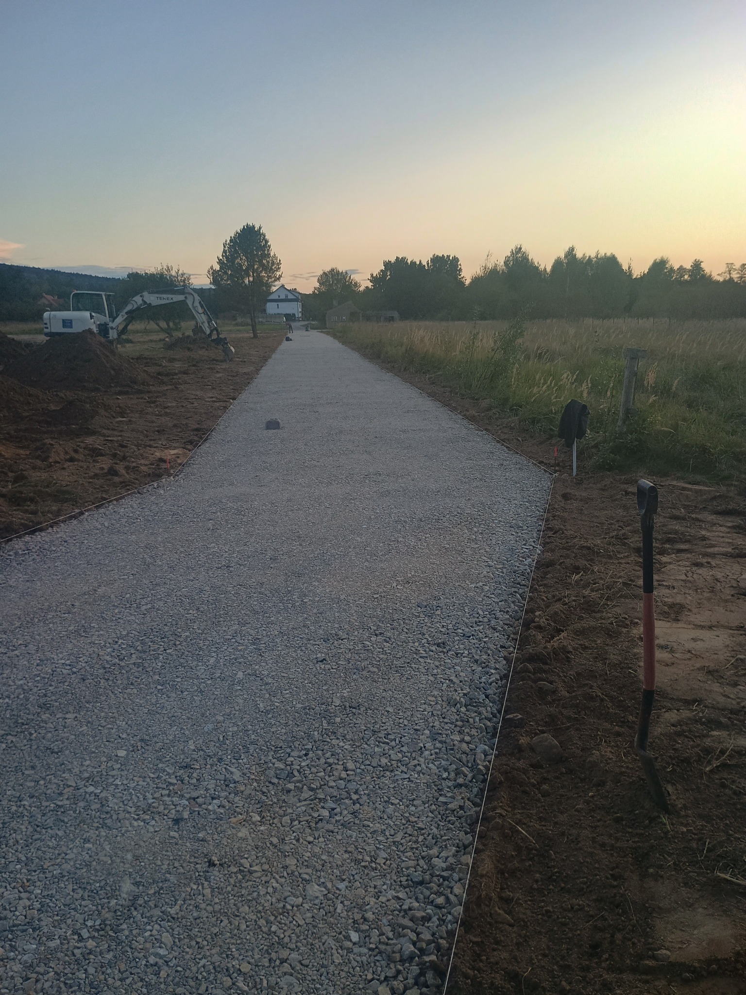 Utwardzona żwirowa droga w perspektywie, z wykoparką w tle, wczesnym rankiem. Widoczne szpadle i wytyczone granice, sugerujące świeżo zakończone prace.