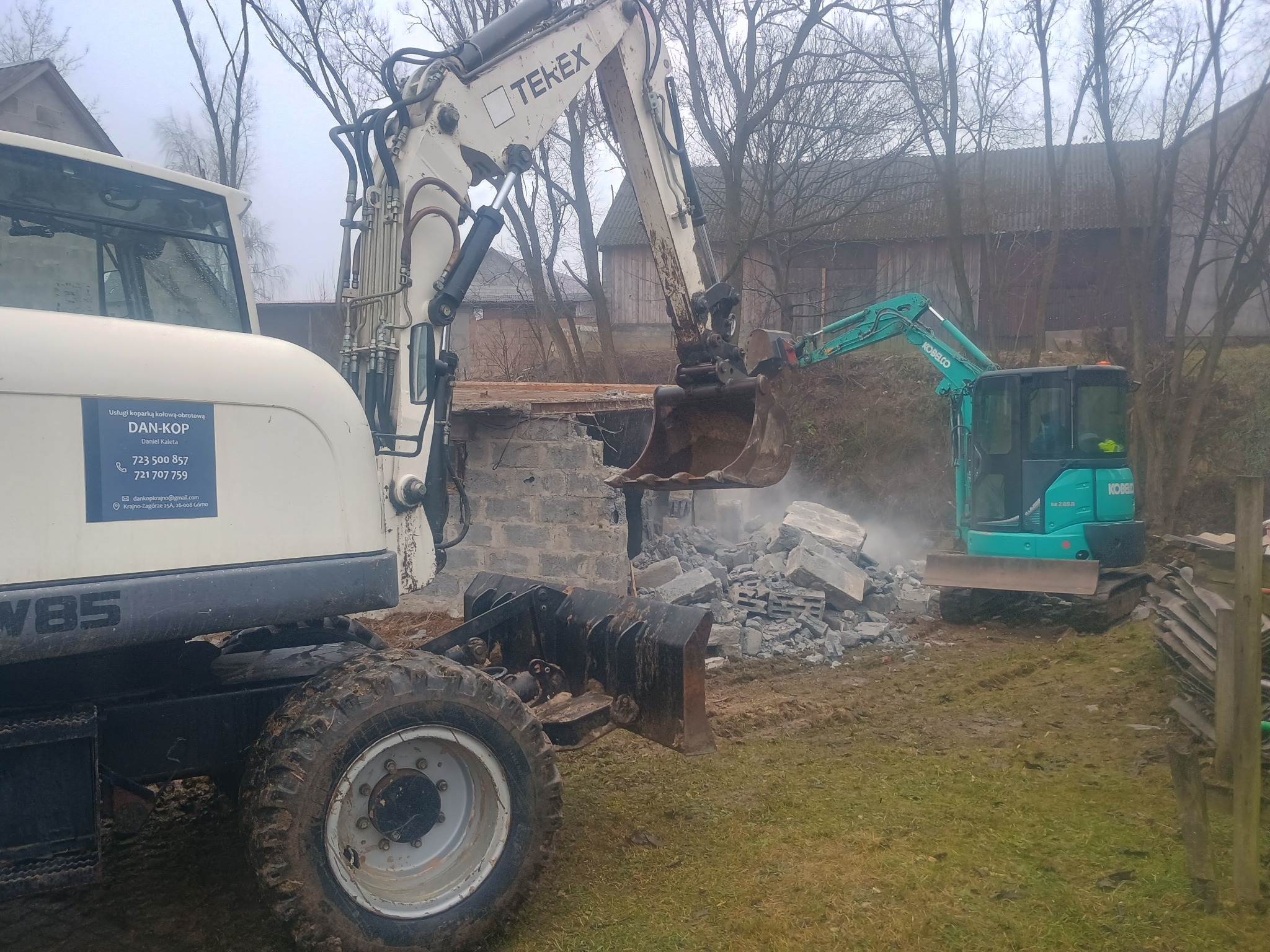 Dwie koparki, biała Tekex i turkusowa Kobelco, podczas rozbiórki murowanej konstrukcji na zewnątrz. Widoczne gruzy i błotniste podłoże. Na białej koparce logo firmy Dan-Kop.