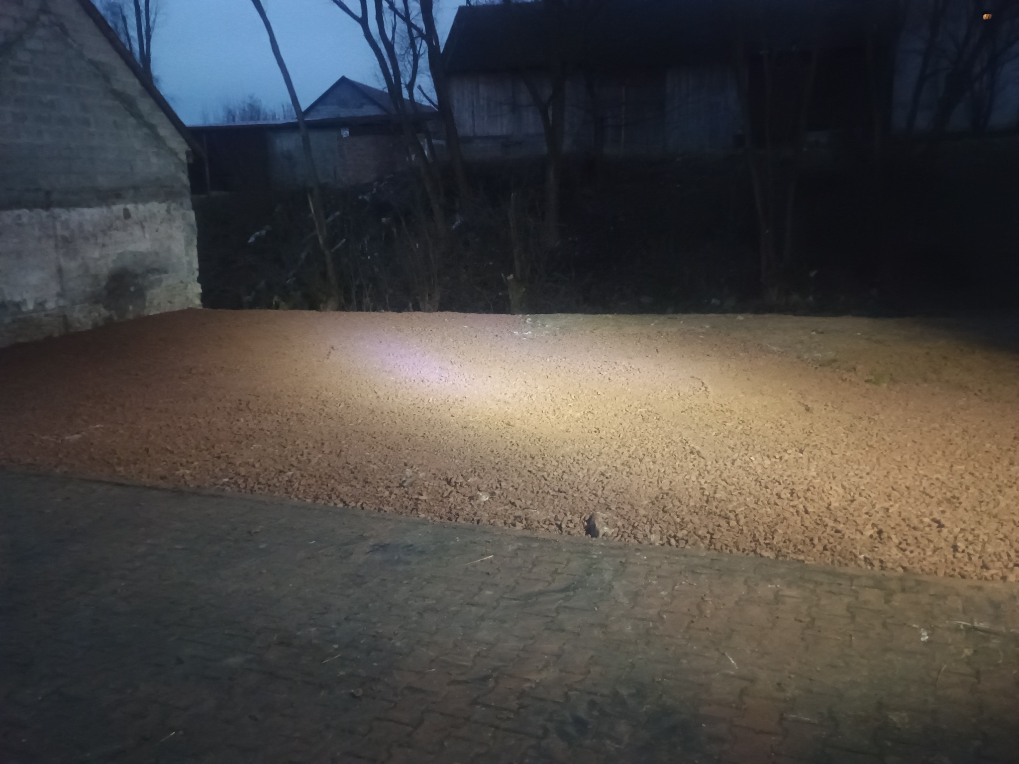 Ustabilizowany teren pod budowę, z widoczną granicą utwardzonej powierzchni i kostką brukową. W tle budynki, oświetlenie wieczorne.