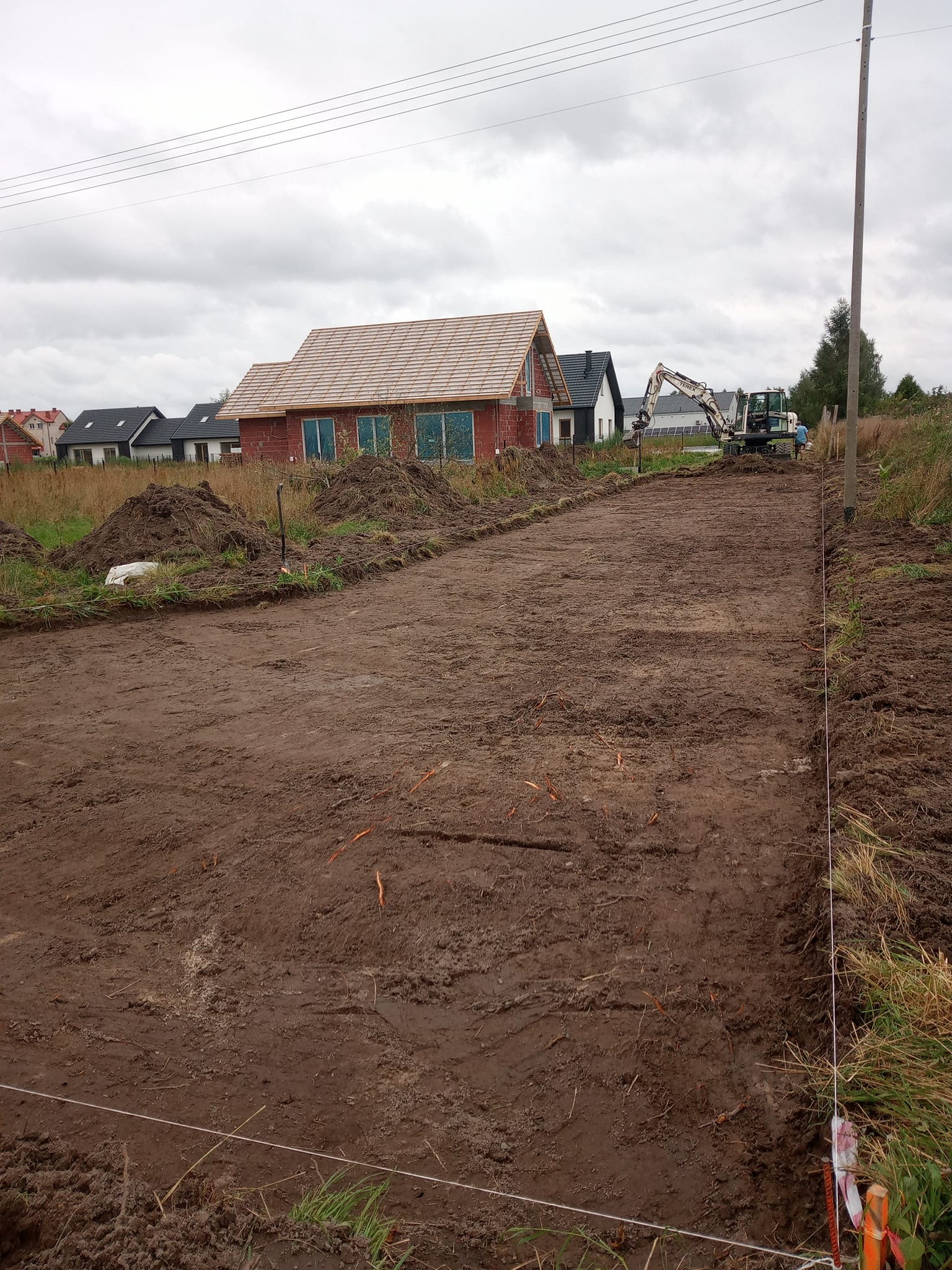 Przygotowany teren pod budowę domu jednorodzinnego z widocznym wykopem i pracującą koparką w tle, w pochmurny dzień. Wytyczone granice działki.
