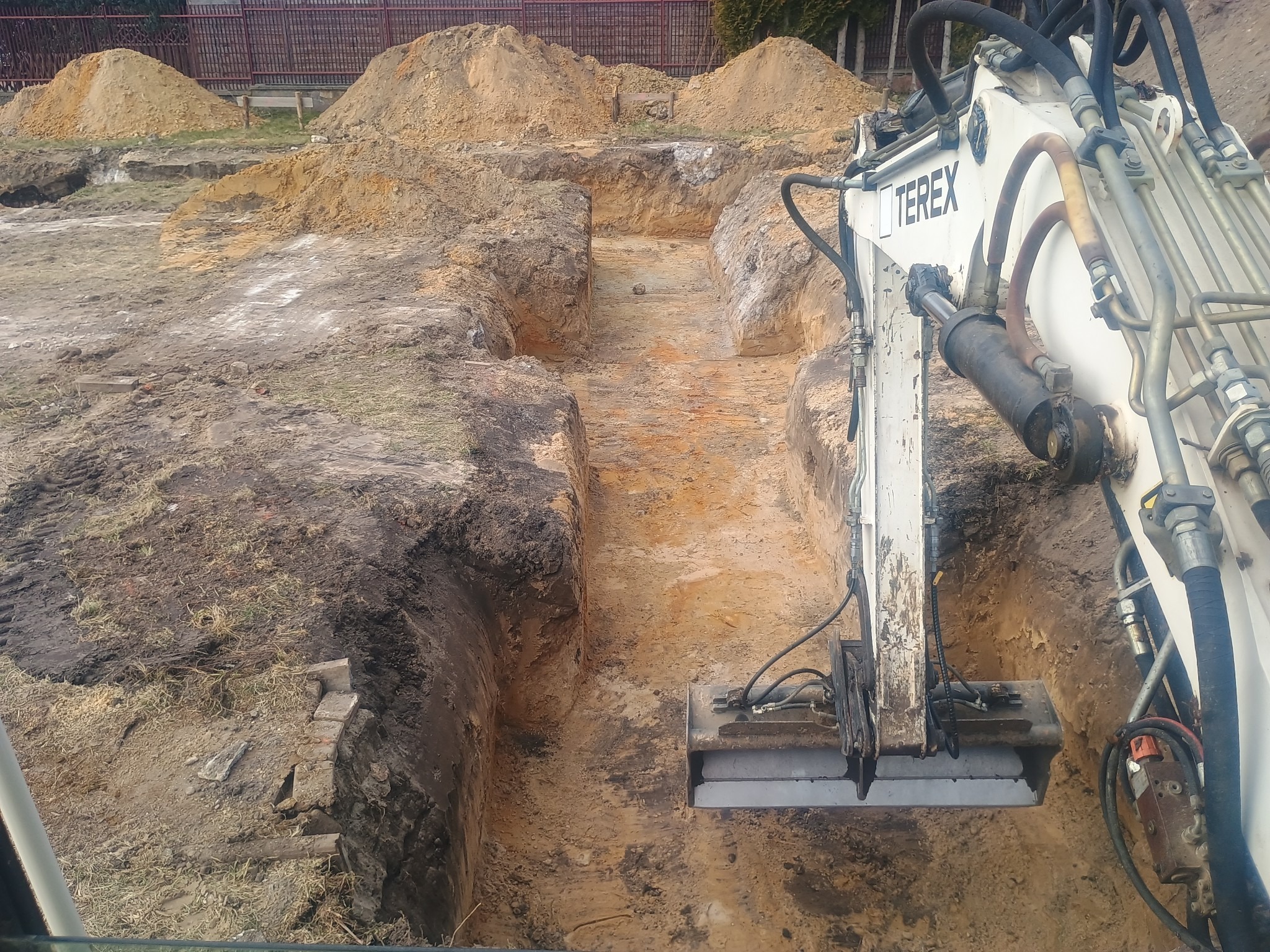Wykop pod fundamenty z użyciem białej koparki Terex. Widoczne wykopane rowy i pryzmy ziemi w tle, ogrodzenie z siatki.