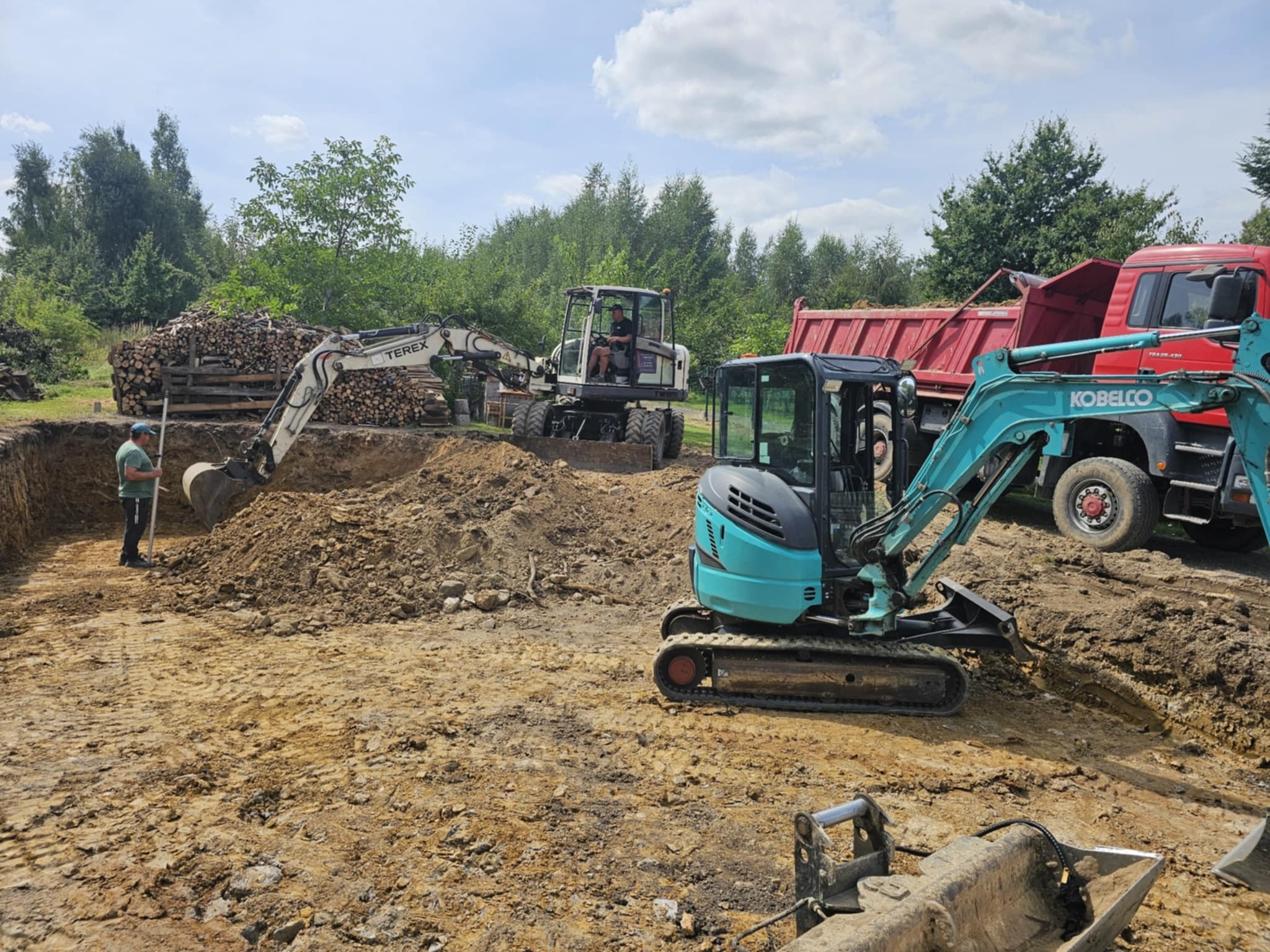 Szeroki kadr na wykop z pracującymi maszynami: koparki Terex i Kobelco, wywrotka. W tle stos drewna. Jeden z pracowników nadzoruje prace ziemne.
