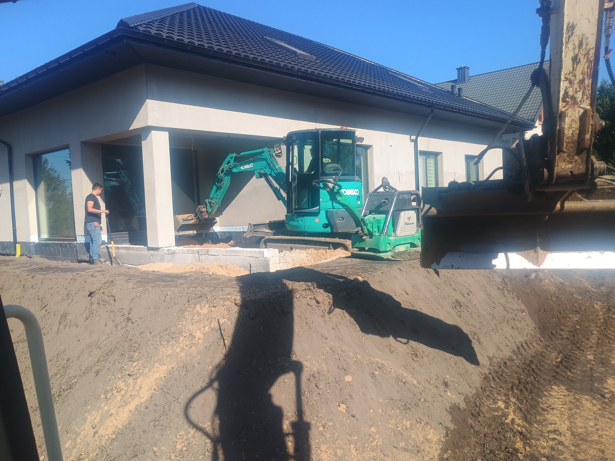 Zielona mini koparka Kobelco na placu budowy domu jednorodzinnego. Widoczny wykop pod fundamenty i pracownik z telefonem. Słoneczny dzień.