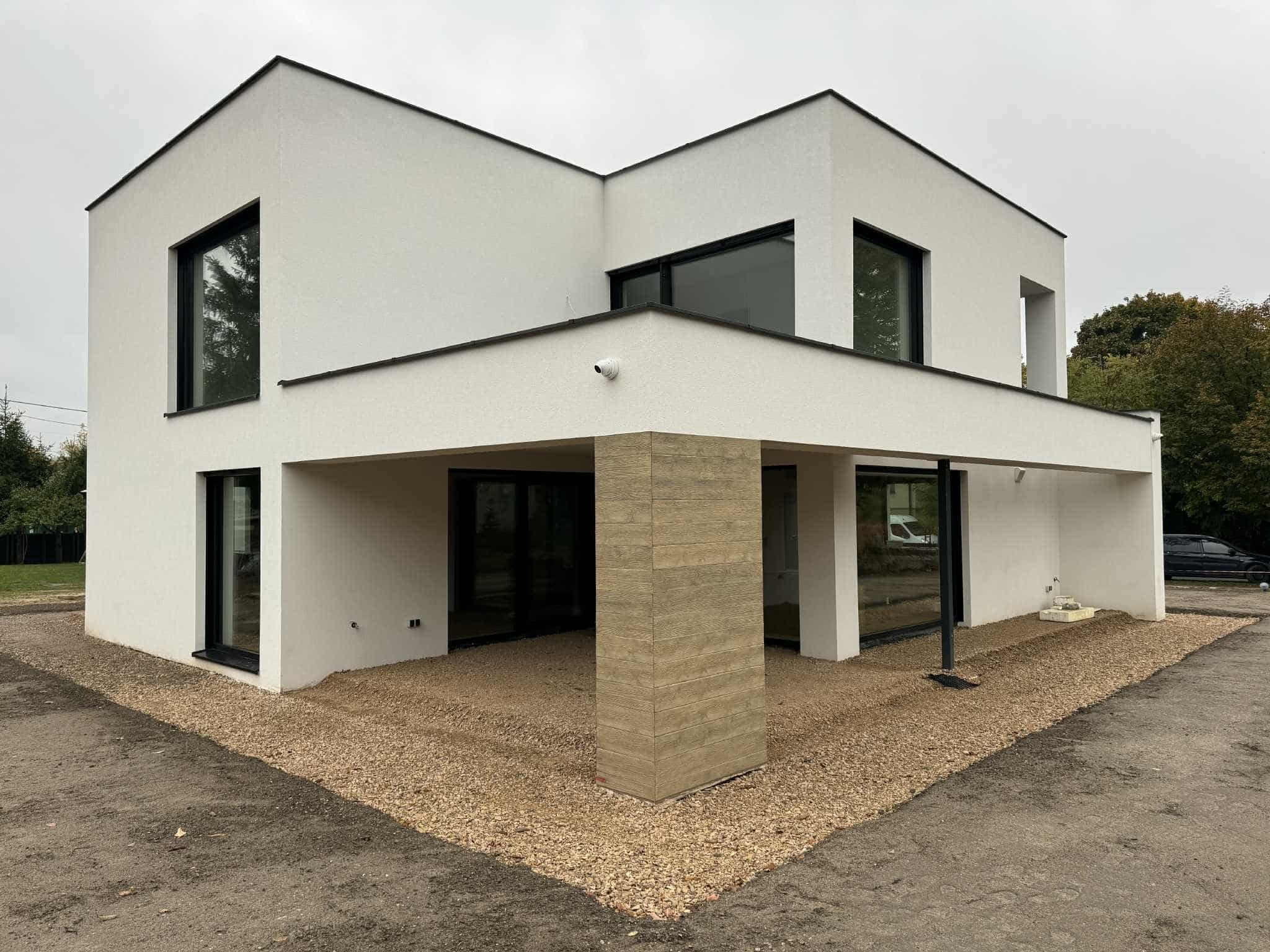 Nowoczesny, biały dom z płaskim dachem i dużymi oknami, otoczony żwirem. Widoczna kolumna z imitacją drewna i fragment czarnego samochodu w tle.
