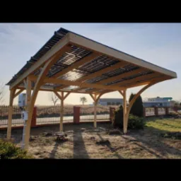 Drewniana wiata z panelami słonecznymi na dachu, widok z zewnątrz, w tle ogrodzenie z ceglanymi słupkami i zabudowania.