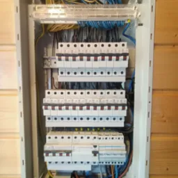 Rozdzielnica elektryczna z widocznymi bezpiecznikami, przewodami w kolorach niebieskim, ż&oacute;łtym i czarnym, zamontowana na ścianie z boazerii.