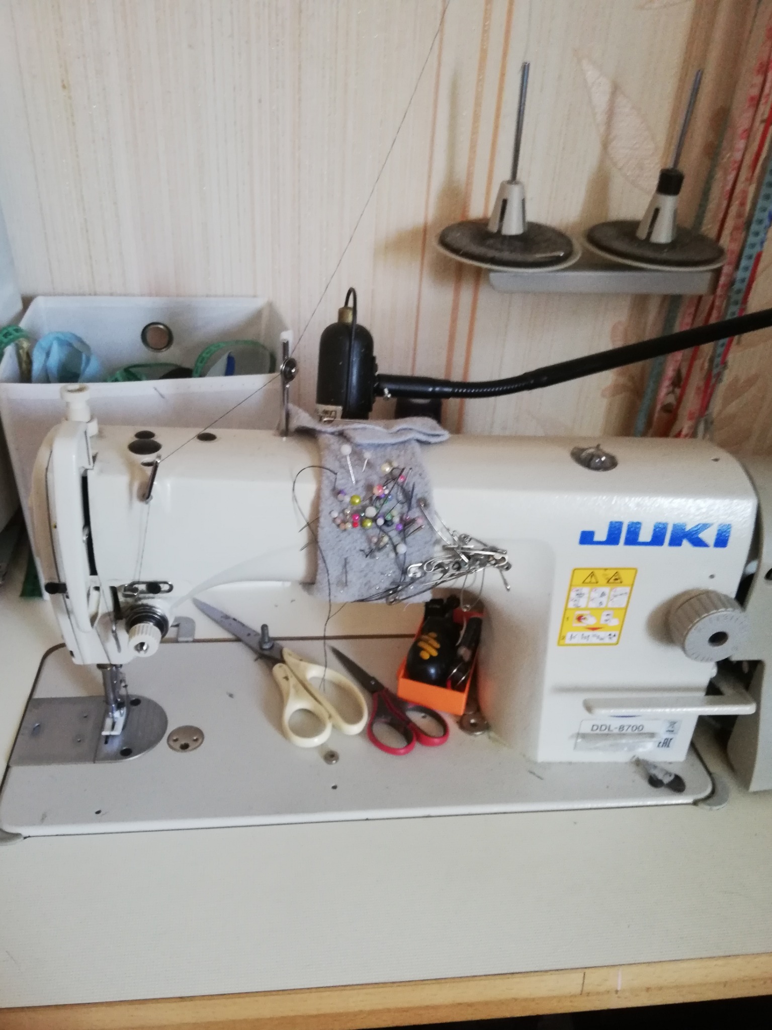 Maszyna do szycia JUKI DDL-8700 z przypiętymi szpilkami do szarej tkaniny, obok nożyczki i pojemnik z akcesoriami, w tle widoczne szpulki nici