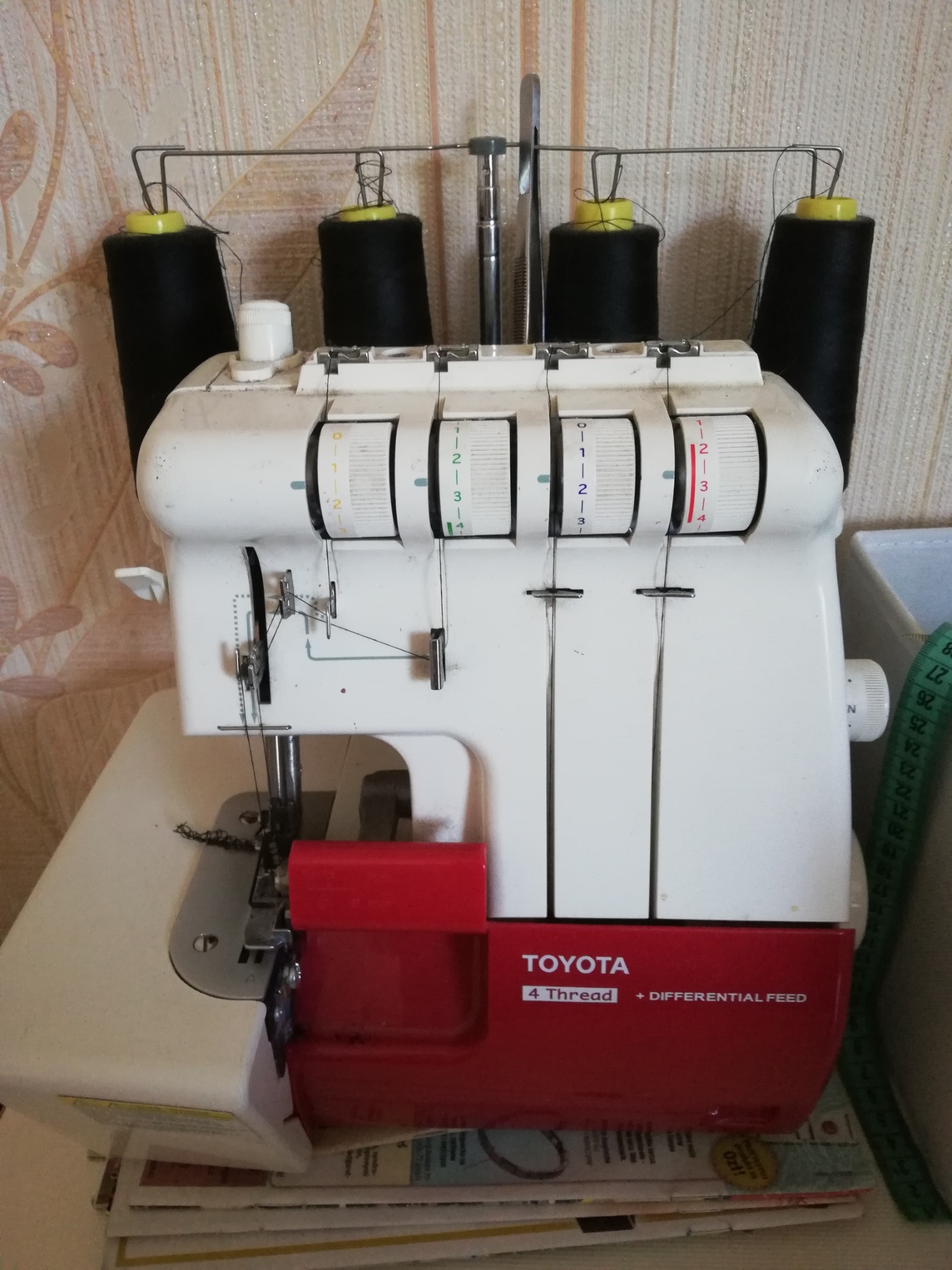 Overlock Toyota z czterema czarnymi szpulkami nici, ustawieniami regulacji i czerwonym pojemnikiem na ścinki, stojący na stosie gazet.