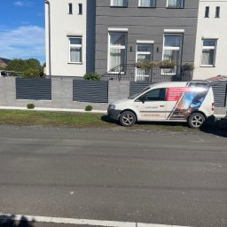 KAM SMART SP. Z O.O. - Nowoczesne ogrodzenie panelowe z szarego betonu i poziomych, grafitowych przęseł, z zaparkowanym samochodem firmy świadczącej usługi spawalnicze.