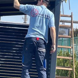 KAM SMART SP. Z O.O. - Pracownik w firmowej koszulce montuje metalową konstrukcję z poziomych profili. W tle drabina i zieleń. Widok z dołu, oświetlenie dzienne.