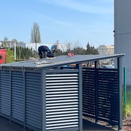 KAM SMART SP. Z O.O. - Montaż metalowej konstrukcji z żaluzjowymi ścianami i skośnym dachem przez pracownika w niebieskiej koszulce, na tle zieleni i budynku.