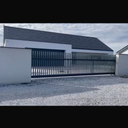 KAM SMART SP. Z O.O. - Nowoczesna, przesuwna brama wjazdowa z pionowych, metalowych profili, na tle domu z szarą dachówką i jasną elewacją. Podłoże wysypane drobnym, szarym żwirem.