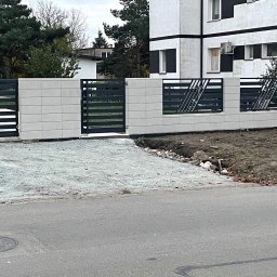 KAM SMART SP. Z O.O. - Nowoczesna brama wjazdowa z poziomymi przęsłami i elementami ukośnymi, osadzona na murze z jasnych, prostokątnych bloków. Widok z ulicy, dom w tle.