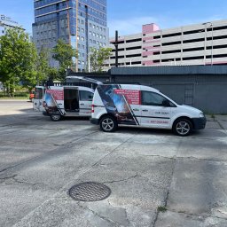 KAM SMART SP. Z O.O. - Dwa białe busy firmy KAM SMART z grafiką spawania i numerem telefonu na tle miejskiego krajobrazu z budynkami i parkingiem. Jeden bus z otwartymi drzwiami.