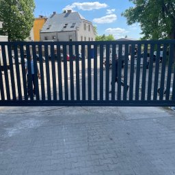 KAM SMART SP. Z O.O. - Nowoczesna, przesuwna brama wjazdowa z pionowych, grafitowych profili, na tle brukowanej nawierzchni i zabudowań miejskich w słoneczny dzień.