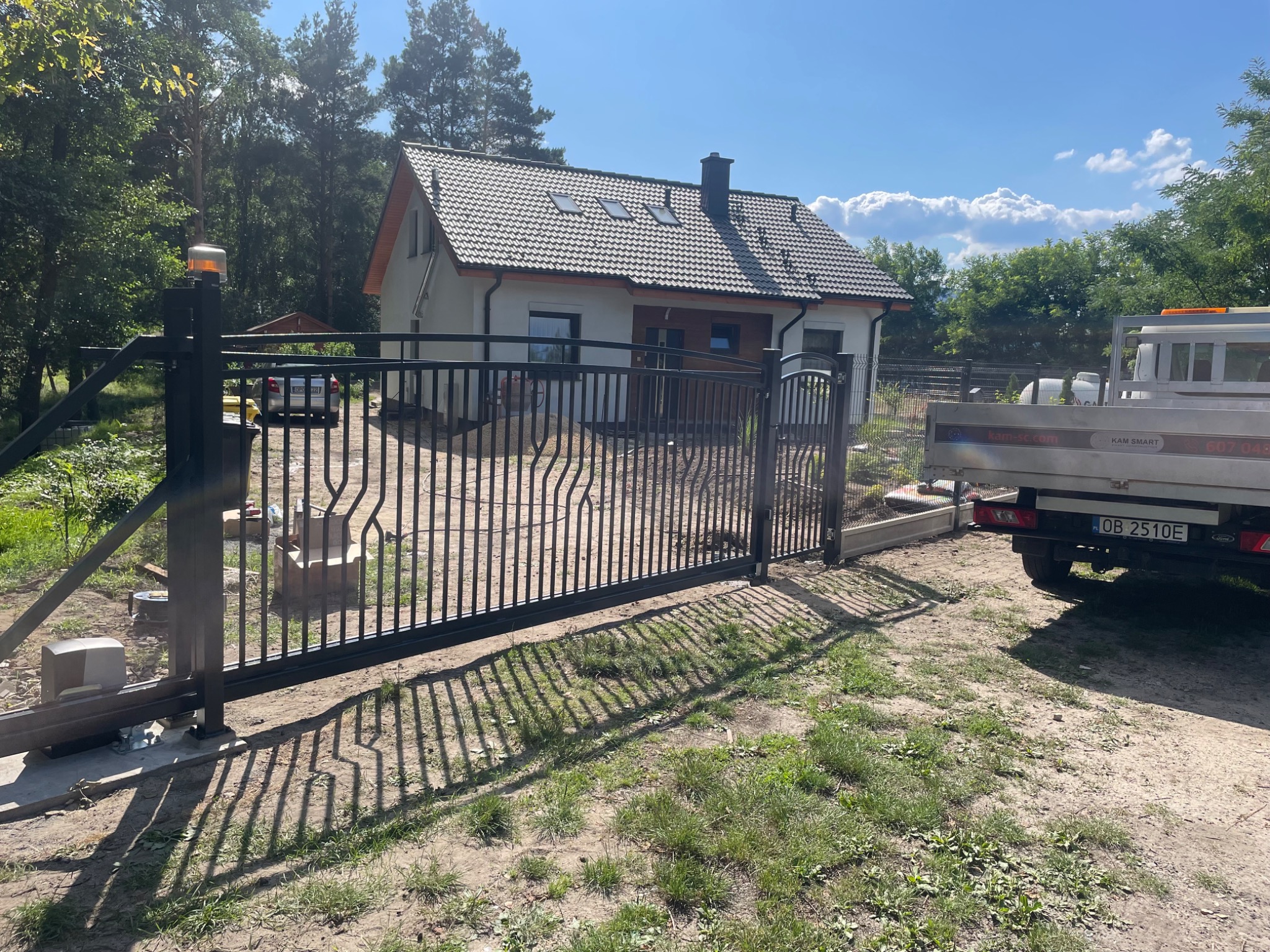 Automatyczna brama przesuwna z czarnego metalu, z widocznym napędem i lampą ostrzegawczą, na tle domu jednorodzinnego i lasu, z zaparkowanym samochodem dostawczym w pobliżu.