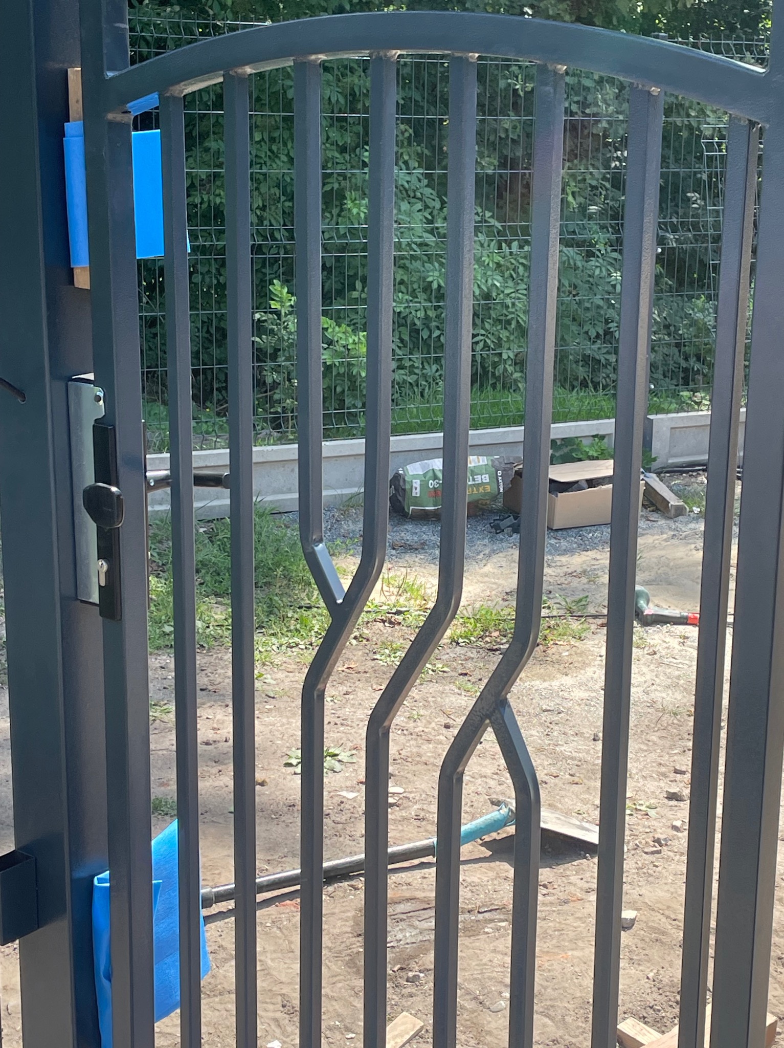 Szara metalowa brama wjazdowa z ozdobnymi pionowymi prętami, widoczny zamek i elementy montażowe, w tle teren budowy z workiem cementu.