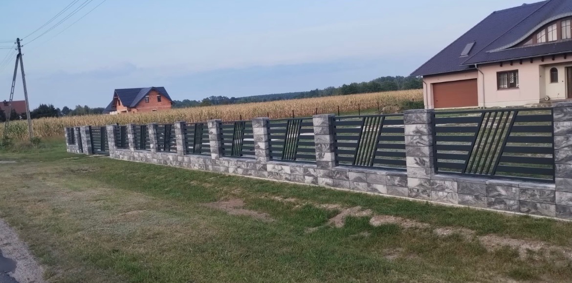 Nowoczesne ogrodzenie z szarej kostki łupanej i metalowych paneli z ukośnymi elementami, na tle pola kukurydzy i domów.