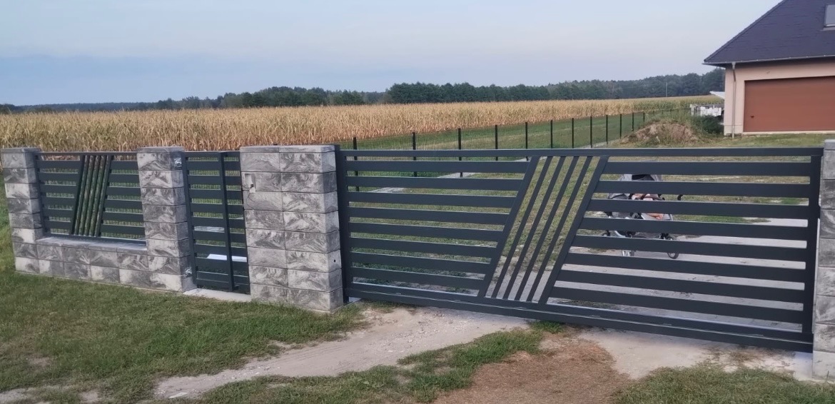 Nowoczesna brama wjazdowa przesuwna z poziomych profili w kolorze grafitowym, z ukośnymi elementami dekoracyjnymi, osadzona na słupkach z szarej kostki, na tle pola kukurydzy i domu.