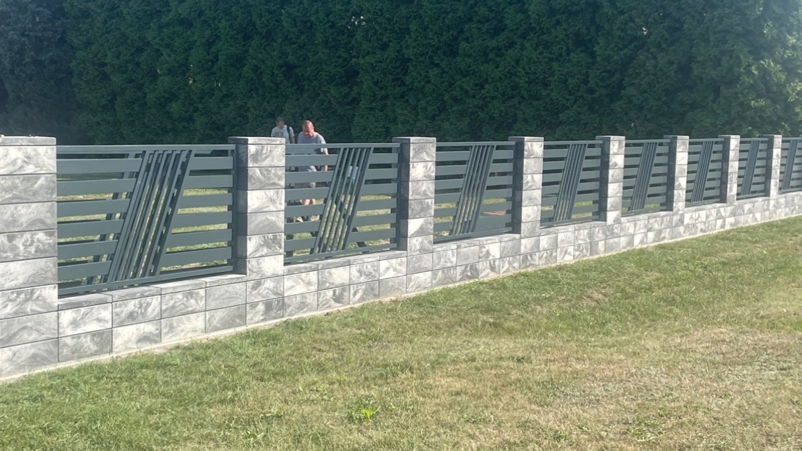 Nowoczesne ogrodzenie panelowe z betonowych bloczków w odcieniach szarości i metalowych przęseł z ukośnymi elementami, na tle zielonego trawnika i żywopłotu, z dwoma osobami w tle.
