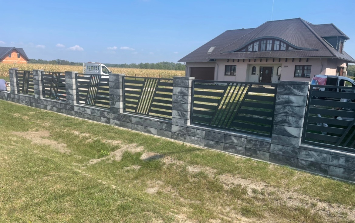 Nowoczesne ogrodzenie panelowe z szarego kamienia łupanego i czarnych, poziomych przęseł z ukośnymi wstawkami, na tle domu z brązowym dachem i zielonego pola kukurydzy.