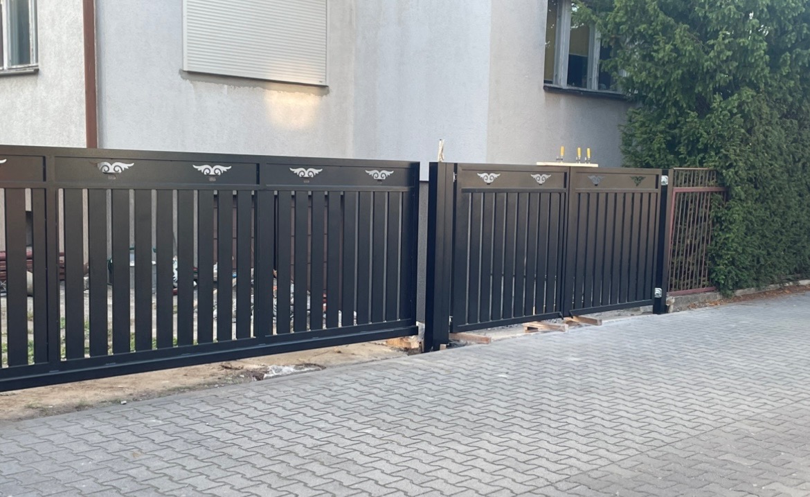Nowoczesna, czarna brama wjazdowa z pionowymi szczeblami i ozdobnymi, ażurowymi motywami na górnej belce, zamontowana na posesji z kostki brukowej, widoczny budynek w tle.