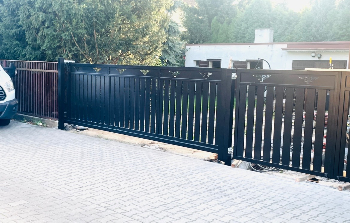 Czarna, przesuwna brama wjazdowa z dekoracyjnymi elementami na tle kostki brukowej i zieleni.