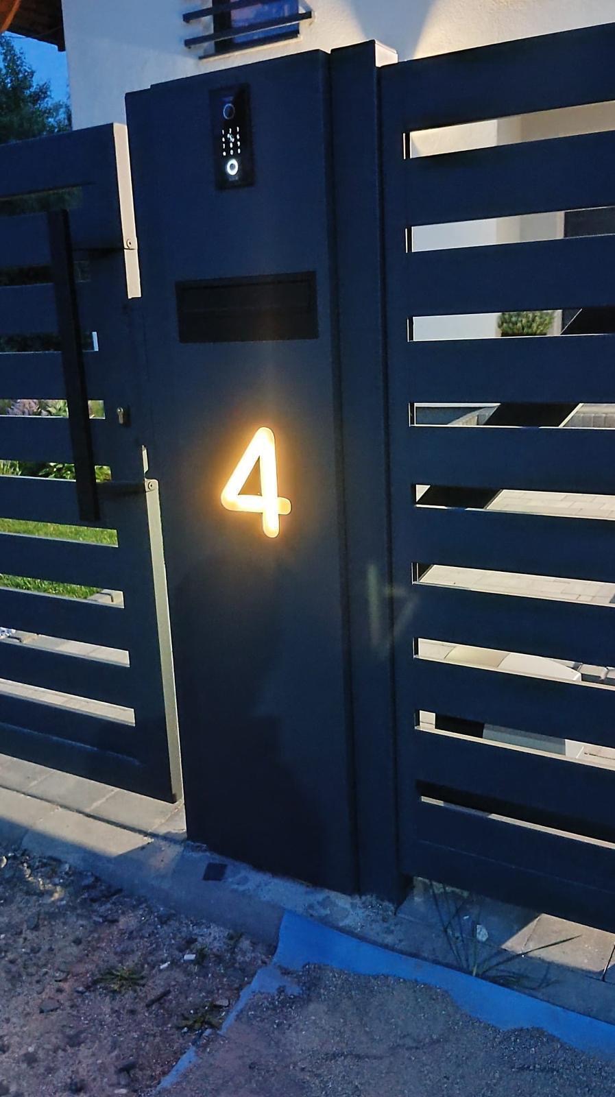 Nowoczesna, grafitowa brama wjazdowa z podświetlanym numerem domu '4' i domofonem, widziana z bliska, prezentuje minimalistyczny design i funkcjonalność.