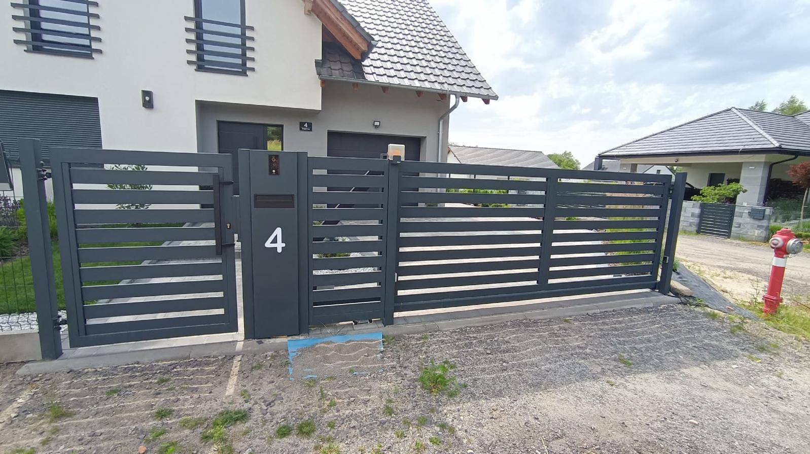 Nowoczesna, pozioma brama wjazdowa w kolorze grafitowym z numerem domu '4', na tle domu z szarą elewacją i dachem z dachówki. Czerwony hydrant w tle.