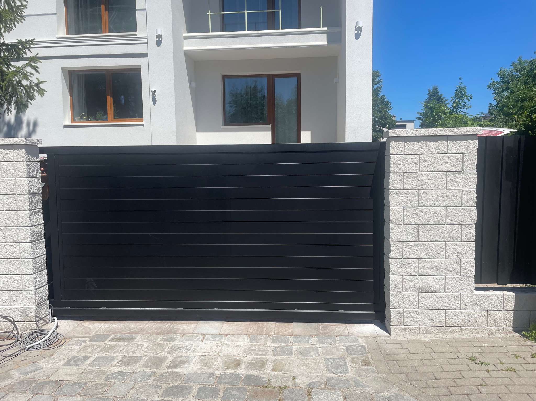 Nowoczesna, pozioma brama wjazdowa w kolorze antracytu, osadzona między dwoma kamiennymi słupkami. Na pierwszym planie kostka brukowa.