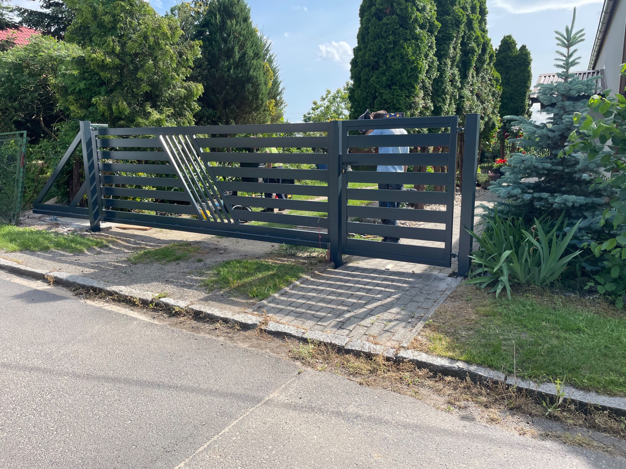 Nowoczesna, szara brama wjazdowa z poziomymi szczeblami i ukośnymi wzmocnieniami, częściowo zamontowana, na tle zieleni i błękitnego nieba. Widoczni pracownicy podczas montażu.