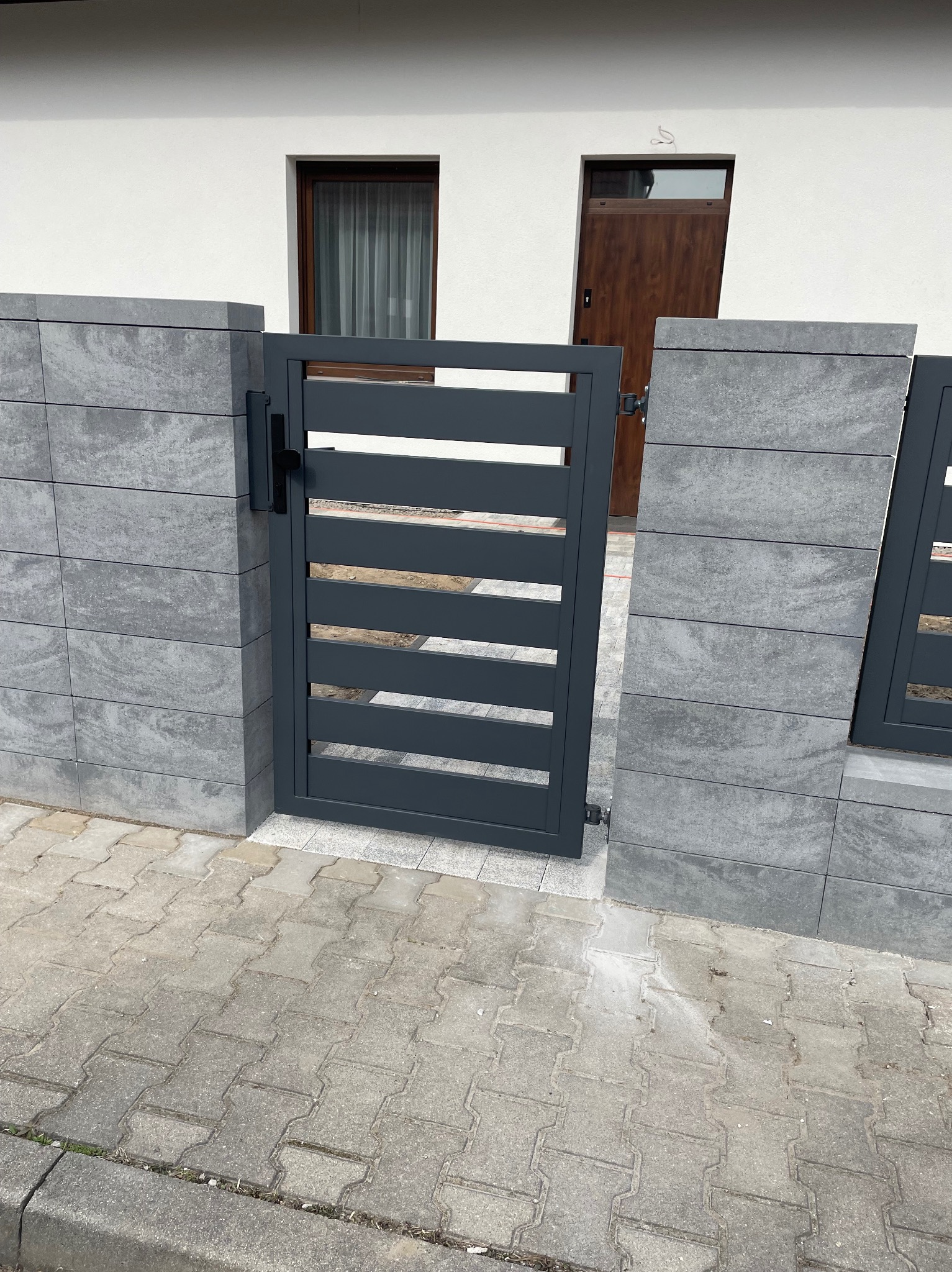 Nowoczesna, antracytowa brama wjazdowa z poziomymi panelami, osadzona między szarymi, betonowymi słupkami. Widoczny fragment domu z oknem i drzwiami.
