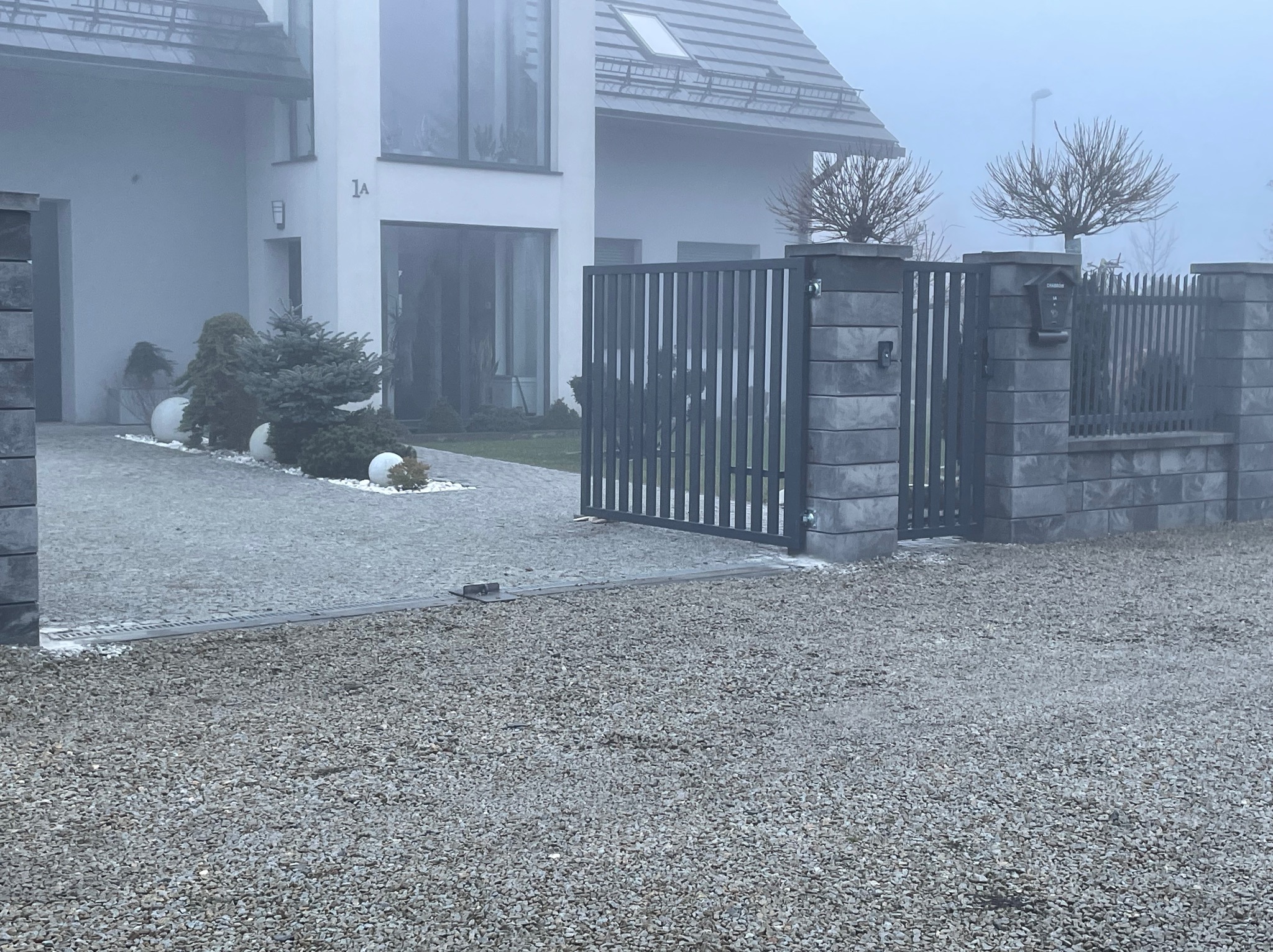 Nowoczesna brama wjazdowa z szarej stali, zintegrowana z ogrodzeniem z betonowych bloczków. Dom w tle, podjazd wysypany żwirem. Mglista pogoda.