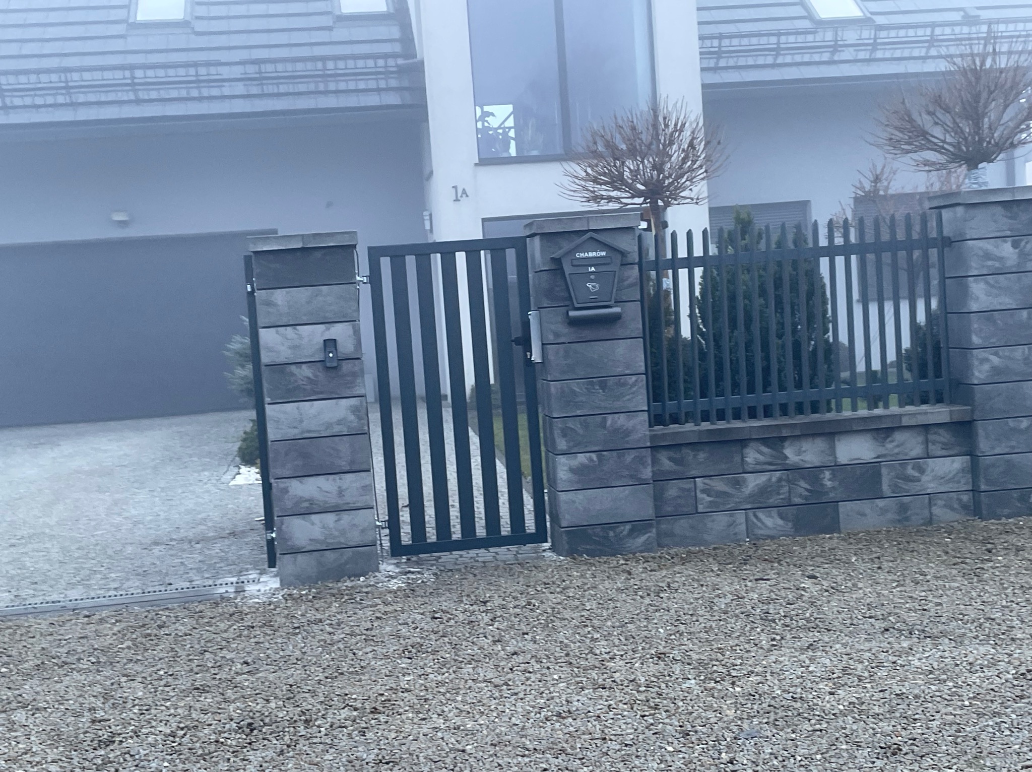 Nowoczesna brama wjazdowa z szarej stali, zintegrowana ze skrzynką pocztową 'CHABROW 1A', na tle domu w mglisty dzień. Minimalistyczny design.