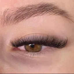 Zbliżenie na oko z przedłużonymi, gęstymi rzęsami w stylu 'kocie oko', widoczna brązowa tęczówka i delikatnie podkreślone brwi.