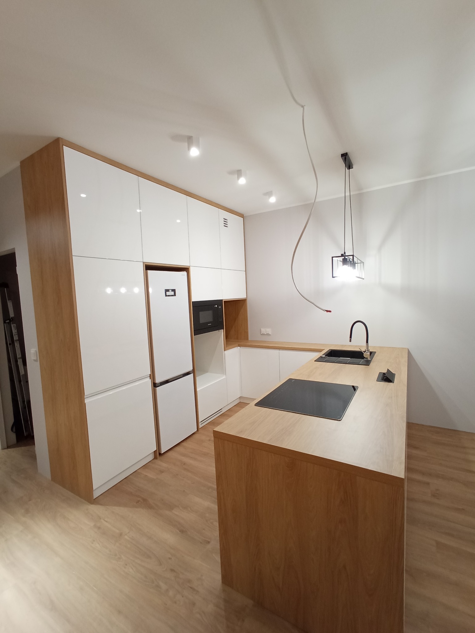 Nowoczesna kuchnia z białymi frontami szafek i drewnianymi elementami wykończeniowymi, wyspa kuchenna z czarnym zlewem i płytą indukcyjną, minimalistyczna lampa wisząca, widoczne instalacje...