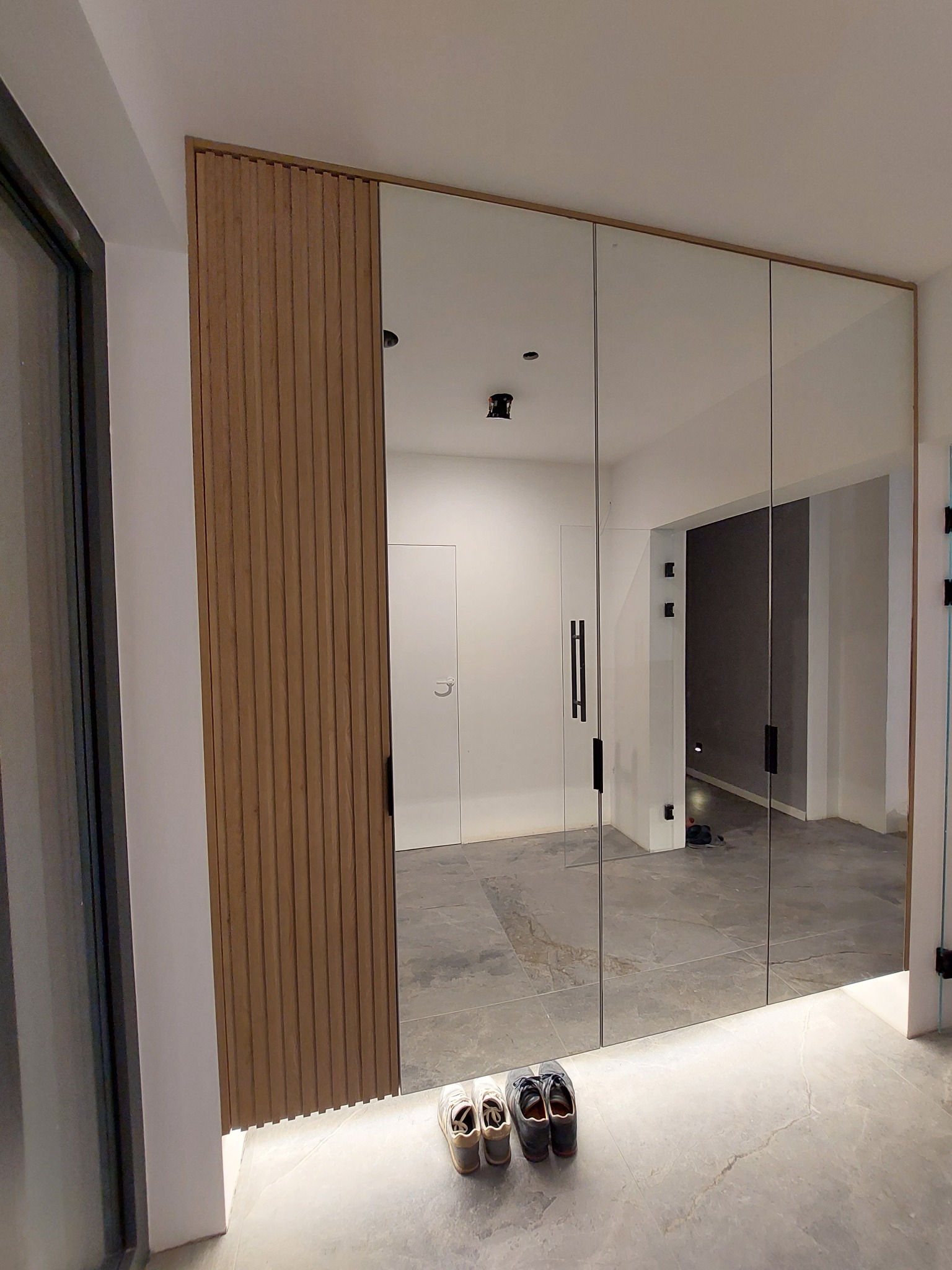 Wielkoformatowe lustro w przedpokoju z drewnianą, lamelową ścianką boczną i podświetleniem LED u dołu, odbijające minimalistyczne wnętrze z szarymi płytkami podłogowymi i trzema parami butów.