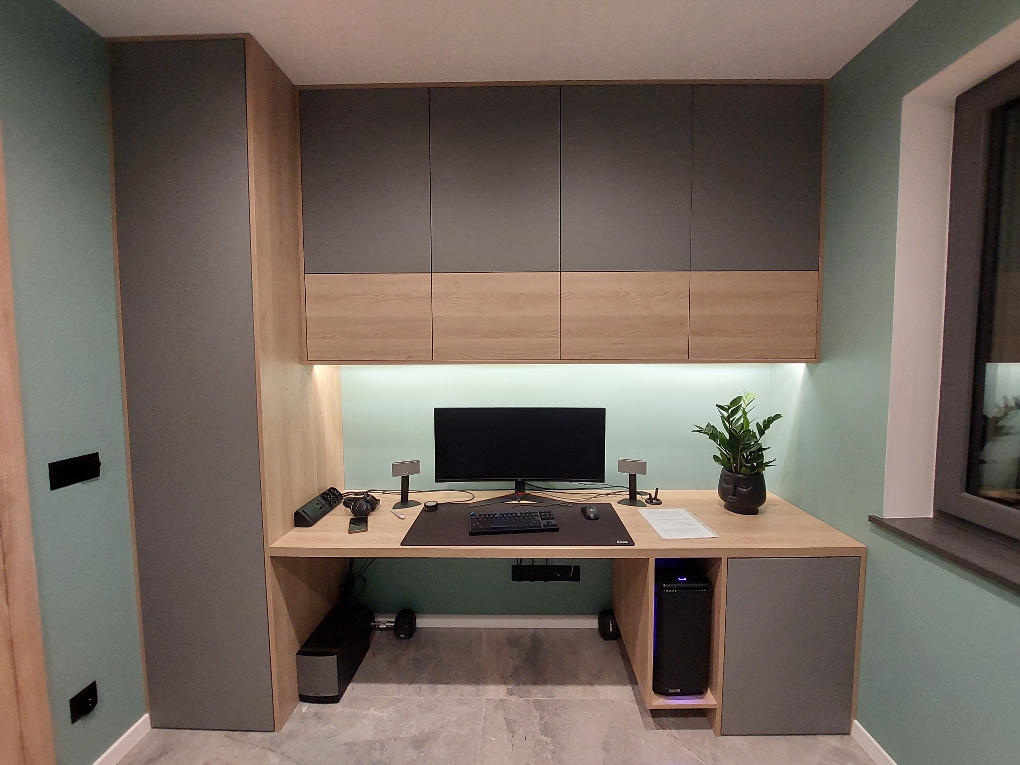 Ergonomiczne stanowisko pracy z blatem z jasnego drewna, szare szafki wiszące i boczne, komputer stacjonarny w obudowie z podświetleniem LED, roślina doniczkowa na biurku, ściana w odcieniu zieleni.
