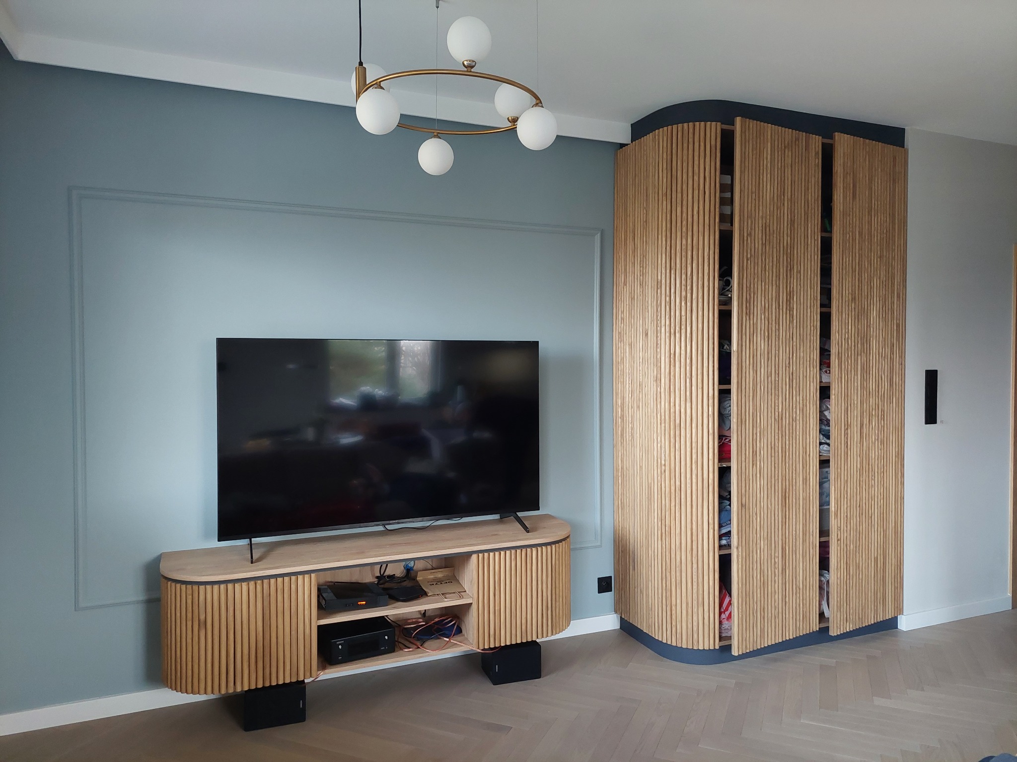 Nowoczesny salon z telewizorem na szafce RTV z lamelami i wysoką szafą z drewnianymi, pionowymi listwami oraz dekoracyjną lampą wiszącą.