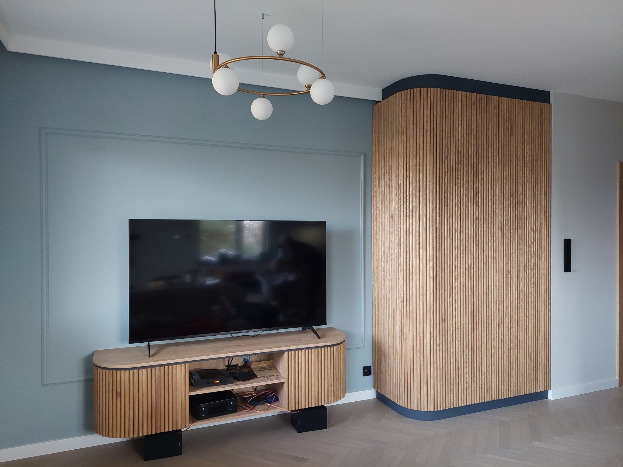Nowoczesny salon z telewizorem na szafce RTV z lamelami, dekoracyjna ściana z pionowych drewnianych listew, oświetlenie sufitowe z kulistymi kloszami, parkiet w jodełkę.