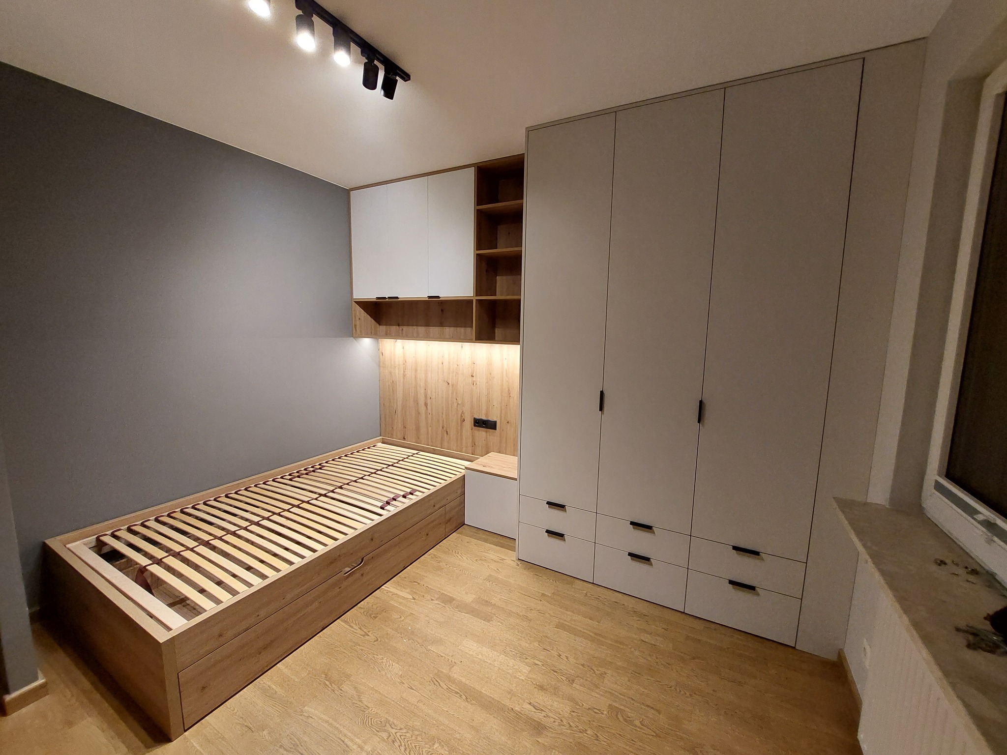 Pokój z łóżkiem drewnianym ze stelażem, szafą wnękową z szufladami i szafkami wiszącymi z drewnianymi półkami, oświetlony reflektorami sufitowymi.