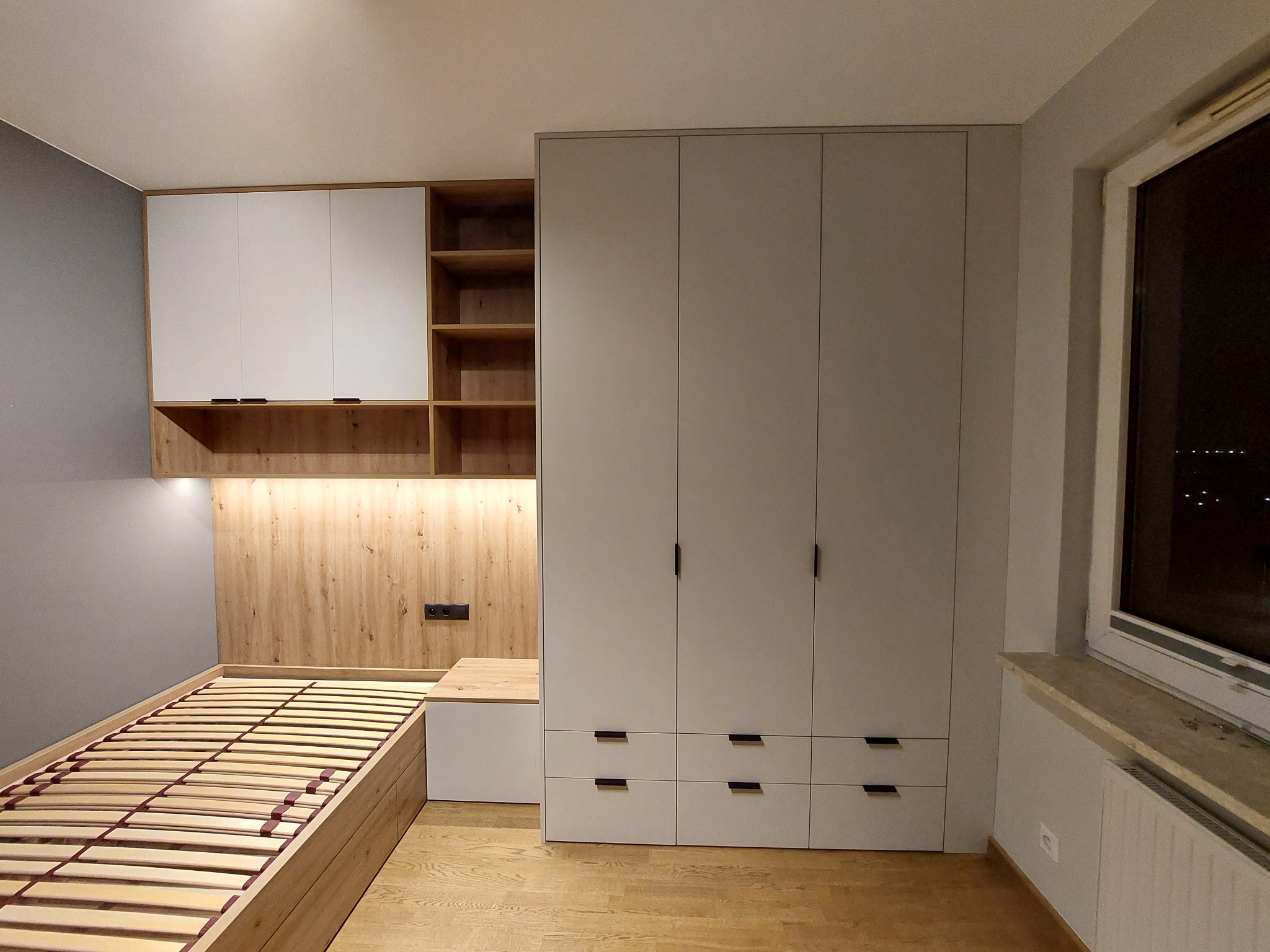 Komplet mebli w pokoju: łóżko z drewnianym stelażem i oświetleniem za wezgłowiem, szafa trzydrzwiowa z szufladami i szafki wiszące z otwartymi półkami.