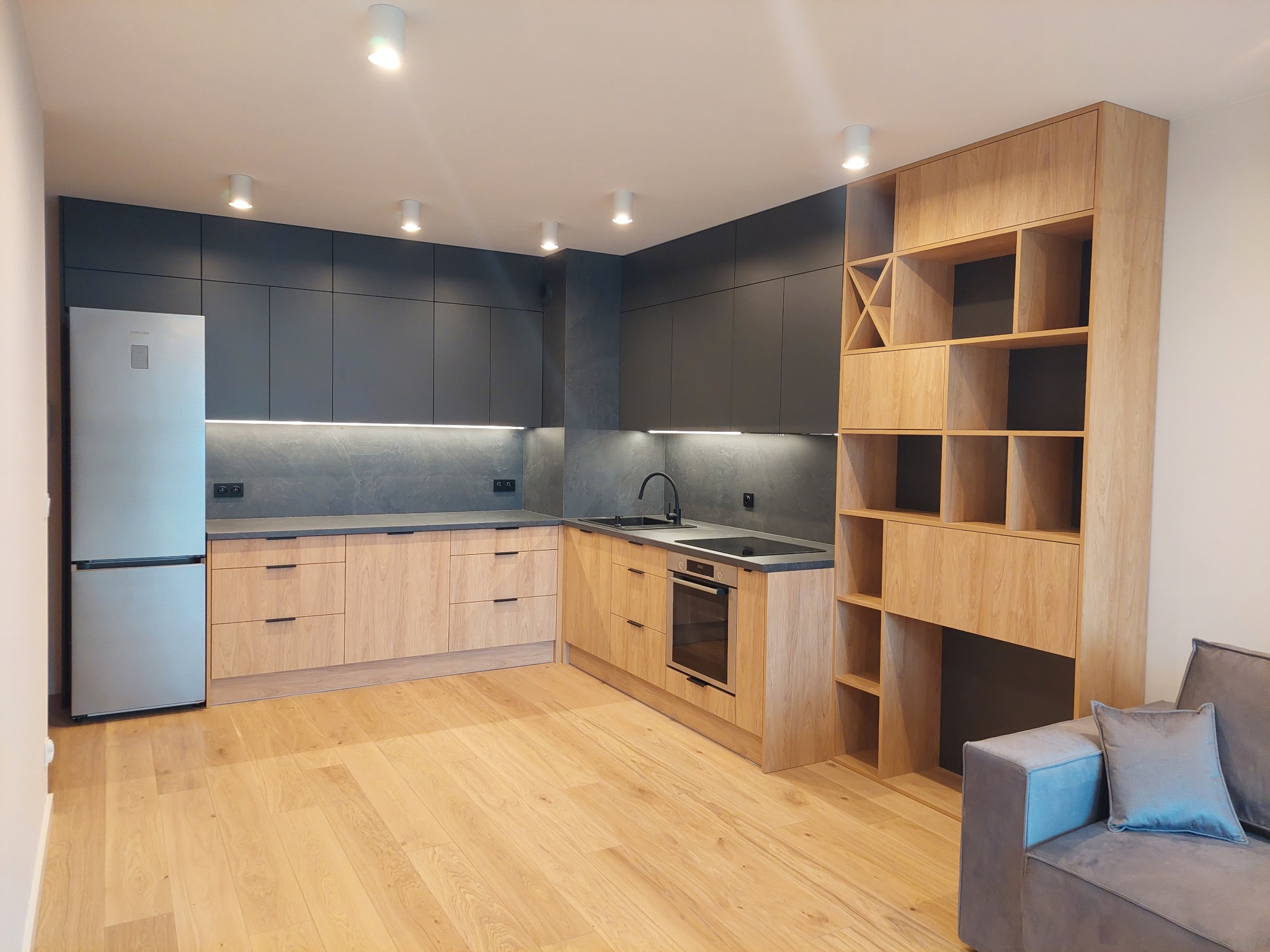 Nowoczesna kuchnia z drewnianymi frontami szafek dolnych i grafitowymi górnymi, oświetleniem LED pod szafkami, drewnianą zabudową regałową z półkami i miejscem na wino, widok z perspektywy salonu...