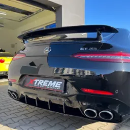 Czarny Mercedes-AMG GT 53 z widocznym logo Xtreme Auto Spa & Detailing na tylnej klapie, zaparkowany na kostce brukowej przed garażem z żółtym Corvette w tle.