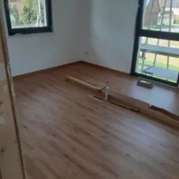 Pokój w trakcie prac wykończeniowych z brązowymi panelami podłogowymi, kartonem po panelach, tubą silikonu i listwami przypodłogowymi, widok z niskiej perspektywy.