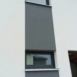 Fasada budynku z dwoma oknami w czarnych ramach, pomalowana w dwóch odcieniach szarości z wyraźną fakturą.