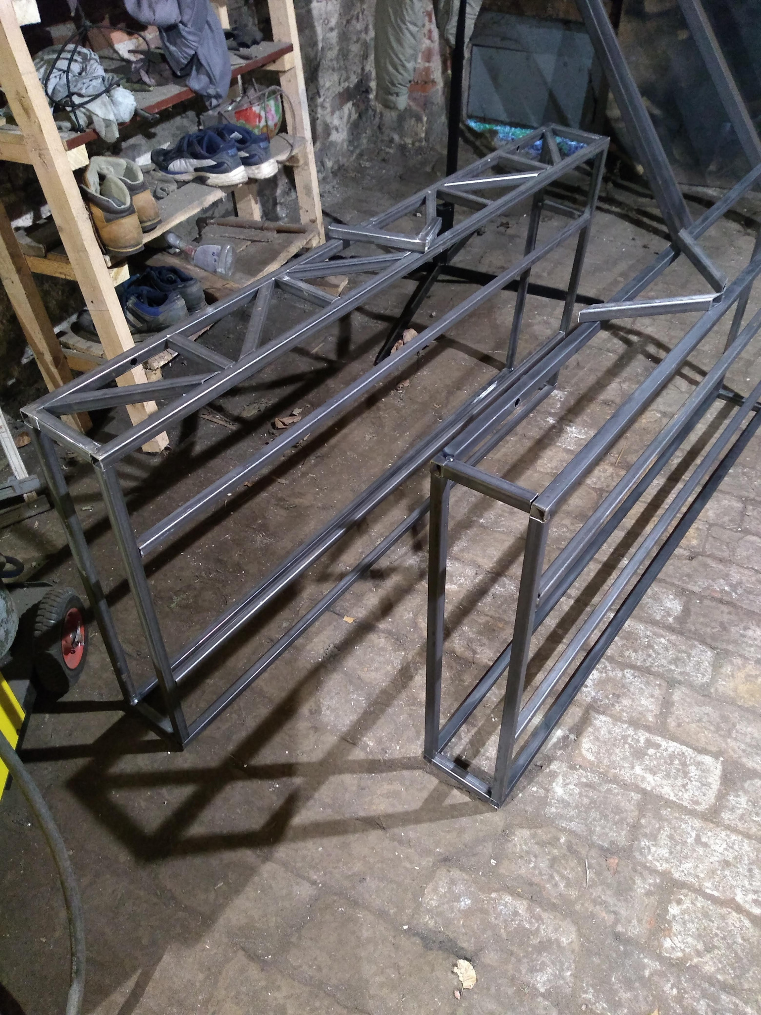 Metalowa konstrukcja z profili kwadratowych, spawana, w surowym stanie, leżąca na ceglanej podłodze warsztatu, widoczny regał z obuwiem w tle.