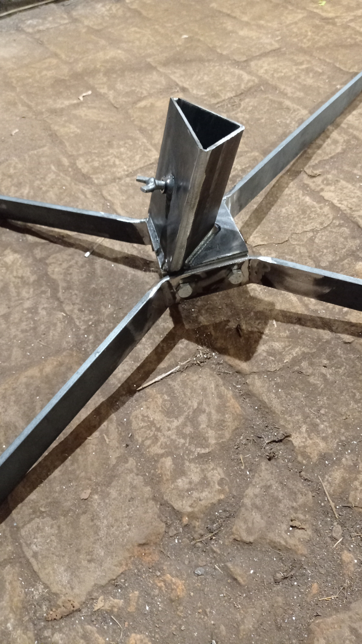 Metalowy stojak z czterema ramionami i kwadratowym otworem na górze, wykonany z profili spawanych, widoczne ślady po spawaniu, umieszczony na brukowanej powierzchni.