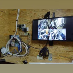 Ekran monitora prezentuje widok z kamer monitoringu w sklepie odzieżowym, obok widoczne kable, zasilacze i lampa na ścianie z płyty OSB.