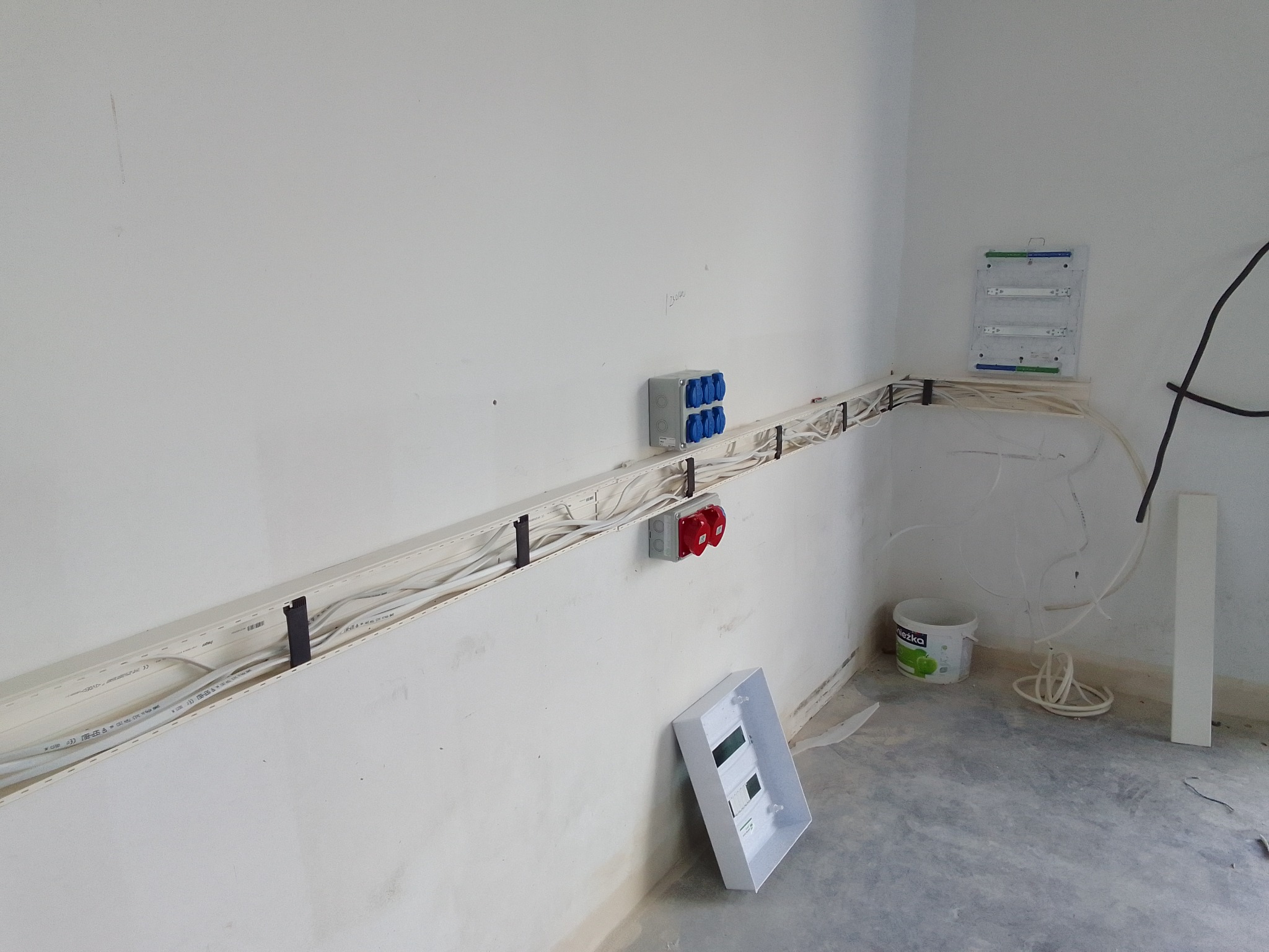 Wnętrze w trakcie prac elektrycznych: widoczne kable w korytkach, gniazda siłowe, puszka rozdzielcza i skrzynka z bezpiecznikami, ściany w trakcie wykończenia, surowa podłoga.